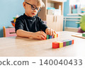 Preschool child with glasses focused on stacking colorful building blocks on a wooden table in a bright classroom with educational toys in the background 140040433