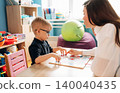 Preschool child with glasses engaged in a learning activity with female teacher at a colorful classroom table, educational materials and toys visible in the background 140040435