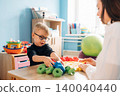 Preschool child with glasses engaged in play with colorful geometric shapes alongside a teacher in a bright classroom filled with educational materials and toys 140040440