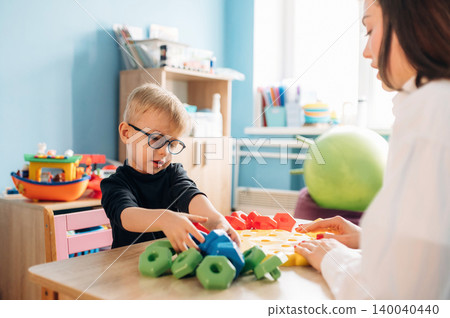 Preschool child with glasses engaged in play with colorful geometric shapes alongside a teacher in a bright classroom filled with educational materials and toys Preschool child with glasses engaged in play with colorful geometric shapes alongside a teacher in a bright classroom filled with educational materials and toys 140040440