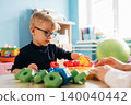 Preschool child with glasses playing with colorful building blocks on a table in a bright classroom, teacher's hand visible guiding the activity 140040442