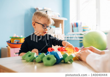 Preschool child with glasses engaged in developmental play with colorful building blocks on a wooden table in a bright classroom environment Preschool child with glasses engaged in developmental play with colorful building blocks on a wooden table in a bright classroom environment 140040444