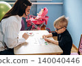 Preschool child with glasses engaged in a learning activity with a female teacher at a wooden table in a colorful classroom with toys in the background 140040446