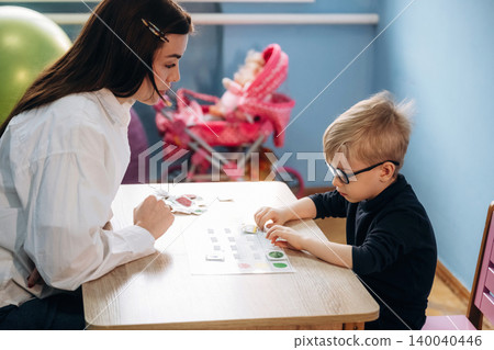 Preschool child with glasses engaged in a learning activity with a female teacher at a wooden table in a colorful classroom with toys in the background Preschool child with glasses engaged in a learning activity with a female teacher at a wooden table in a colorful classroom with toys in the background 140040446