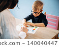 Preschool child wearing glasses engages with teacher at wooden table, colorful learning materials visible, pink chair in background, educational setting for developmental activities 140040447