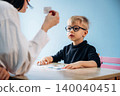 Preschool child with glasses engaged in learning activity with teacher holding flashcard at a table in a bright classroom setting with blue wall background 140040451