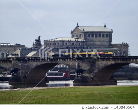Yellow tram and river ferry passing under bridge, commuters, boat traffic and cloudy urban backdrop 140040504