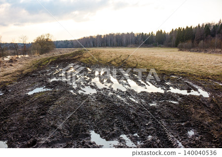Gnarly, slushy mud track, riddled with puddles and tire ruts, traversing a desolate field towards a distant forest line under an overcast sky Gnarly, slushy mud track, riddled with puddles and tire ruts, traversing a desolate field towards a distant forest line under an overcast sky 140040536