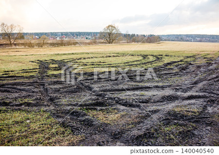 Churned field, deep tire tracks marring the landscape. Dark, wet mud contrasts with patches of green grass under an overcast sky, hinting at rural life 140040540
