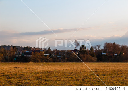 Vast golden field leads to a quiet village of houses and bare trees under a serene, cloudy sky. An idyllic countryside panorama Vast golden field leads to a quiet village of houses and bare trees under a serene, cloudy sky. An idyllic countryside panorama 140040544