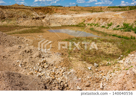 Open pit quarry with a reflective puddle, surrounded by dry, cracked earth and rubble under a blue sky with clouds Open pit quarry with a reflective puddle, surrounded by dry, cracked earth and rubble under a blue sky with clouds 140040556