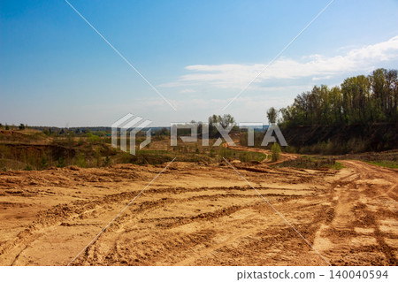 Old sand quarry terrain with numerous vehicle tire tracks. Barren slopes and distant green trees under a bright blue sky, revealing a rugged outdoor landscape Old sand quarry terrain with numerous vehicle tire tracks. Barren slopes and distant green trees under a bright blue sky, revealing a rugged outdoor landscape 140040594