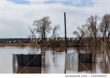 Submerged beach changing cabins during a river flood, with muddy waters overflowing the bank. Bare trees and distant houses visible under a cloudy sky. Nature's power on display Submerged beach changing cabins during a river flood, with muddy waters overflowing the bank. Bare trees and distant houses visible under a cloudy sky. Nature's power on display 140040607
