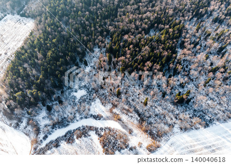 Aerial winter forest view. Evergreen and bare trees blanketed in snow near a frozen creek and snowy fields. Nature's frosty grip Aerial winter forest view. Evergreen and bare trees blanketed in snow near a frozen creek and snowy fields. Nature's frosty grip 140040618