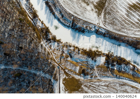 Icy river meandering through snow-covered winter terrain, flanked by a stark forest and frosty fields. A peaceful aerial vista of nature's chill 140040624