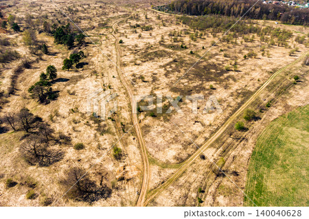 Aerial view of a winding dirt road snaking through dry, sparse spring fields. Bare trees and scattered green patches dot the sun-drenched landscape Aerial view of a winding dirt road snaking through dry, sparse spring fields. Bare trees and scattered green patches dot the sun-drenched landscape 140040628