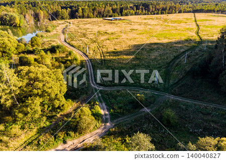 Winding country roads crisscross a sprawling field under golden light, flanked by a serene pond and dense woods, hinting at secluded rural paths Winding country roads crisscross a sprawling field under golden light, flanked by a serene pond and dense woods, hinting at secluded rural paths 140040827