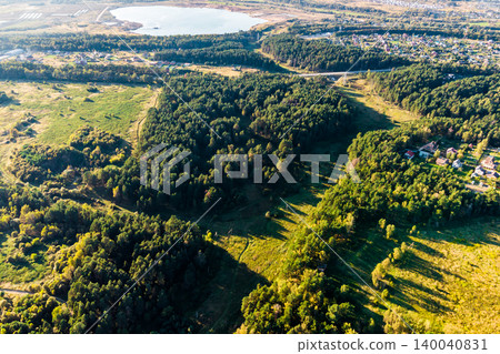 Aerial view of a sprawling green forest, interspersed with sunny fields and a quaint village. A tranquil lake shimmers in the distance, completing this serene natural panorama Aerial view of a sprawling green forest, interspersed with sunny fields and a quaint village. A tranquil lake shimmers in the distance, completing this serene natural panorama 140040831