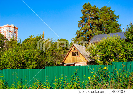 Charming rustic wooden cottage peeks over a vibrant green fence, nestled amidst lush trees. A tall apartment building hints at urban closeness under a clear sky Charming rustic wooden cottage peeks over a vibrant green fence, nestled amidst lush trees. A tall apartment building hints at urban closeness under a clear sky 140040861