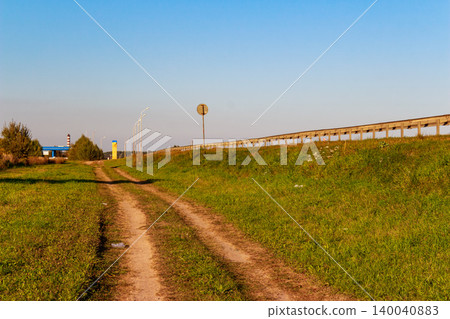 A rustic dirt track meanders beside a modern highway, with a protective guardrail delineating the edge. Distant streetlights and a gas station add hints of development A rustic dirt track meanders beside a modern highway, with a protective guardrail delineating the edge. Distant streetlights and a gas station add hints of development 140040883