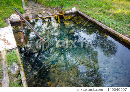 Rustic immersion basin fed by a natural spring, featuring clear, tranquil water vividly reflecting overhead trees and a simple handrail Rustic immersion basin fed by a natural spring, featuring clear, tranquil water vividly reflecting overhead trees and a simple handrail 140040907
