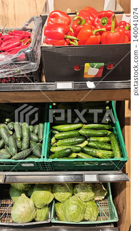 Fresh red bell peppers, cucumbers, and green cabbage displayed in crates on wooden shelves inside a grocery store produce section Fresh red bell peppers, cucumbers, and green cabbage displayed in crates on wooden shelves inside a grocery store produce section 140041074