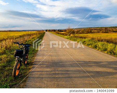 Electric fat bike parked by a long, sunny country road. Golden fields stretch under a dynamic sky, a perfect pit stop on an outdoor jaunt Electric fat bike parked by a long, sunny country road. Golden fields stretch under a dynamic sky, a perfect pit stop on an outdoor jaunt 140041099