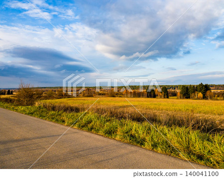 A country lane runs by a golden field with lush green verges. Far-off trees line the horizon under a vast, cloud-strewn sky A country lane runs by a golden field with lush green verges. Far-off trees line the horizon under a vast, cloud-strewn sky 140041100