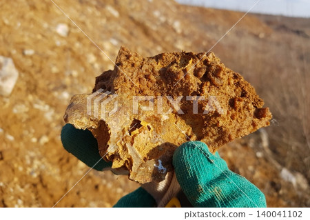 A gloved hand cradles a rough, earthy piece of opaque chalcedony on flint, showcasing its rugged texture against a soft-focus barren landscape under bright sunlight 140041102