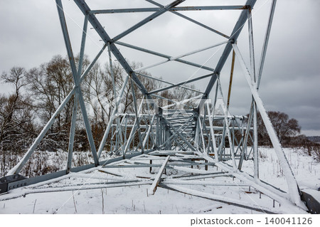 A power line pylon's metallic frame lays covered in fresh snow across a wintry field, awaiting erection. Bare trees dot the cloudy horizon A power line pylon's metallic frame lays covered in fresh snow across a wintry field, awaiting erection. Bare trees dot the cloudy horizon 140041126