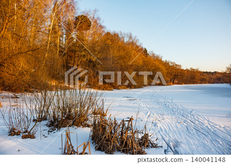 Golden light bathes a serene frozen river, its snowy expanse marked by ski tracks. Bare trees line the bank, adding a warm glow to the tranquil winter landscape Golden light bathes a serene frozen river, its snowy expanse marked by ski tracks. Bare trees line the bank, adding a warm glow to the tranquil winter landscape 140041148