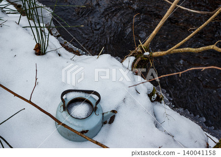 An old, light blue enameled kettle lies abandoned in fresh snow near a dark stream, surrounded by bare winter branches. A forlorn scene of discarded utility 140041158