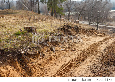 Gritty dirt track with distinct tire marks ascends a bare, erosion-prone slope. Scattered tree stumps hint at deforestation, beneath a dull, late autumn sky Gritty dirt track with distinct tire marks ascends a bare, erosion-prone slope. Scattered tree stumps hint at deforestation, beneath a dull, late autumn sky 140041168