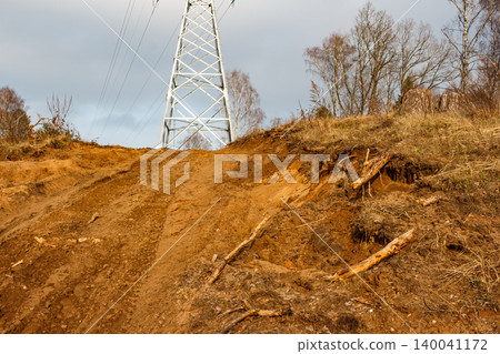 Damaged raw earth slope, scarred by tracks and exposed roots, rises to a stark power pylon under a moody sky, showing environmental disruption Damaged raw earth slope, scarred by tracks and exposed roots, rises to a stark power pylon under a moody sky, showing environmental disruption 140041172