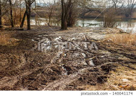 Mucky, rutted ground filled with tire tracks and puddles, showcasing a challenging off-road terrain. Bare trees line the distant river Mucky, rutted ground filled with tire tracks and puddles, showcasing a challenging off-road terrain. Bare trees line the distant river 140041174