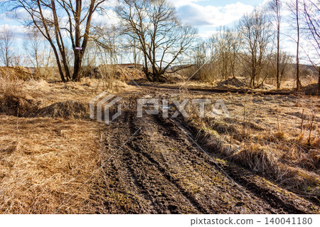 A gnarly, muddy dirt track cuts through withered grass and bare trees under a bright sky, hinting at tough off-road journey A gnarly, muddy dirt track cuts through withered grass and bare trees under a bright sky, hinting at tough off-road journey 140041180