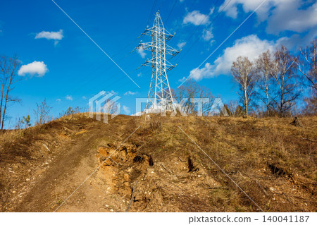 A robust power line pylon stands tall on a dry hillside under a bright blue sky with sparse clouds. Industrial landscape meets nature's raw beauty A robust power line pylon stands tall on a dry hillside under a bright blue sky with sparse clouds. Industrial landscape meets nature's raw beauty 140041187