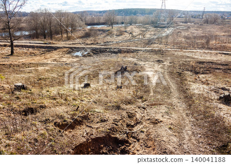 Gnarly dirt paths cut across a raw, parched expanse dotted with old tree stumps and withered scrub. A river and distant woods frame the untamed scene Gnarly dirt paths cut across a raw, parched expanse dotted with old tree stumps and withered scrub. A river and distant woods frame the untamed scene 140041188