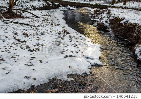 Sparkling winter stream partly frozen, flanked by snowy banks and ice-covered ground. Sunlight glints on flowing water, highlighting nature's serene chill Sparkling winter stream partly frozen, flanked by snowy banks and ice-covered ground. Sunlight glints on flowing water, highlighting nature's serene chill 140041213