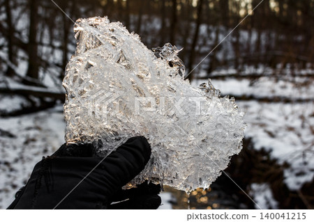 A gloved hand cradles a shimmering piece of ice, showcasing intricate patterns against a blurred wintry forest backdrop. Melting ice drips 140041215