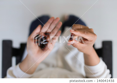 Hands hold two pills, one pink, one green. Person rests in a cozy, softly lit room. Calm demeanor suggests routine or health care. Perfect for wellness, medicine, or health Hands hold two pills, one pink, one green. Person rests in a cozy, softly lit room. Calm demeanor suggests routine or health care. Perfect for wellness, medicine, or health 140041438