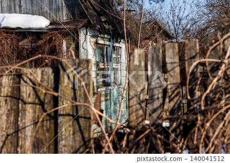 Old wooden dacha cottage with peeling paint behind a rustic weathered fence in early spring, melting snow patches on the roof, abandoned countryside scenery under bright daylight Old wooden dacha cottage with peeling paint behind a rustic weathered fence in early spring, melting snow patches on the roof, abandoned countryside scenery under bright daylight 140041512