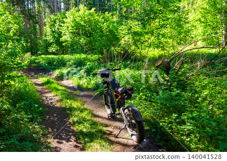 Electric fat bike parked on a narrow forest dirt path surrounded by lush vibrant greenery and summer foliage in a sunny tranquil woodland setting for outdoor recreation adventure 140041528