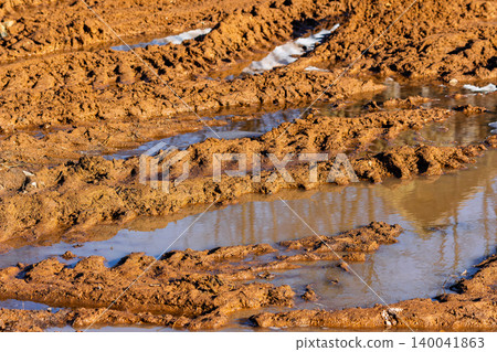 Deep tire tracks carved into thick wet clay soil creating a muddy and messy off-road trail filled with stagnant rainwater puddles after heavy spring rains 140041863