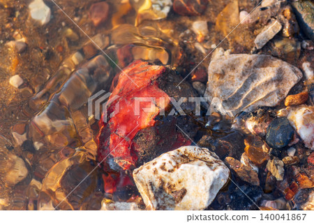 Striking red flint fragment submerged in clear river water, gleaming amongst diverse riverbed stones. Natural beauty and unique geology close-up Striking red flint fragment submerged in clear river water, gleaming amongst diverse riverbed stones. Natural beauty and unique geology close-up 140041867