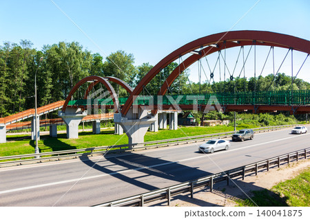 Modern arched pedestrian bridge over a multi-lane highway with moving cars, surrounded by lush green trees on a sunny day providing urban transportation infrastructure Modern arched pedestrian bridge over a multi-lane highway with moving cars, surrounded by lush green trees on a sunny day providing urban transportation infrastructure 140041875