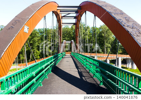 Modern pedestrian arched bridge with orange steel structures and vibrant green railings crossing over a highway, providing a safe walking path Modern pedestrian arched bridge with orange steel structures and vibrant green railings crossing over a highway, providing a safe walking path 140041876