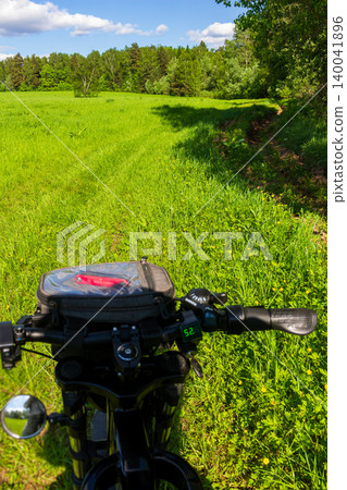 Close-up perspective of an electric bike cockpit featuring a handlebar bag and digital display, navigating through a lush vibrant green meadow on a sunny summer day Close-up perspective of an electric bike cockpit featuring a handlebar bag and digital display, navigating through a lush vibrant green meadow on a sunny summer day 140041896
