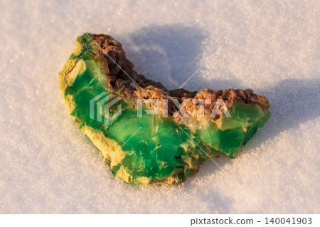 Vibrant green Kazakh chrysoprase specimen, raw and earthy, nestled on a pristine blanket of sparkling white snow, highlighting its natural beauty Vibrant green Kazakh chrysoprase specimen, raw and earthy, nestled on a pristine blanket of sparkling white snow, highlighting its natural beauty 140041903