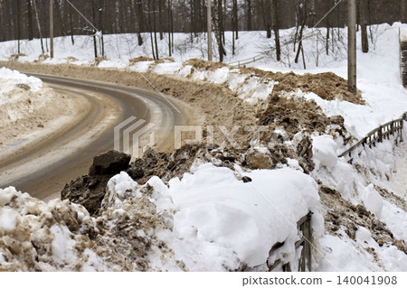 Hefty dirty snowdrifts heap along a winding winter road, cleared by plows. Brown slush and white snow bury the roadside fence, showcasing challenging winter aftermath 140041908